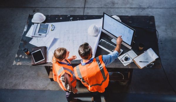 High angle shot of two engineers working together on blueprints while making use of a computer inside of a workshop